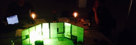 a dim room of gamers gathered around a table. An green light illuminates a Handler's screen and notes.