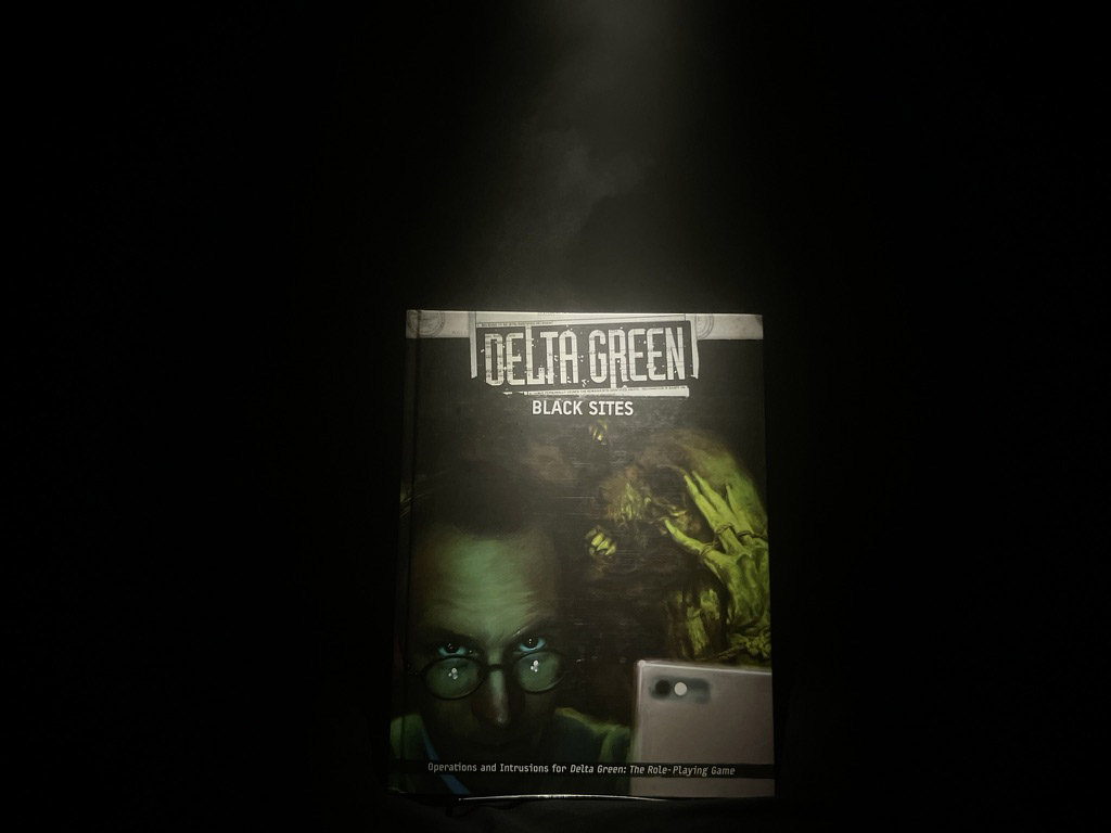 A copy of Delta Green: Black Sites sits in the darkness illuminated by a smoky spotlight from above.